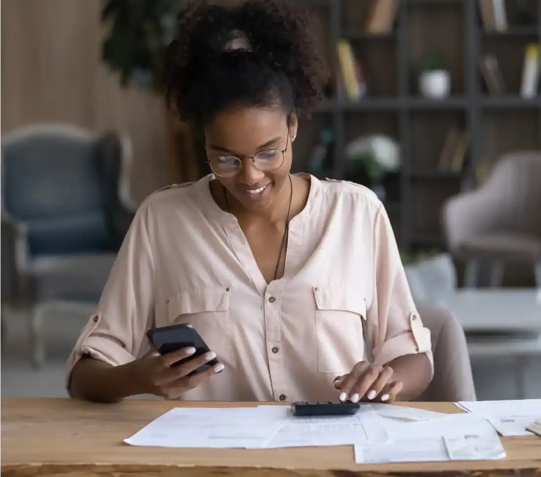 Mulher negra com oculos e cabelos presos sentada em frente a uma mesa com o celular na mão aparentemente fazendo calculos na calculadora.
